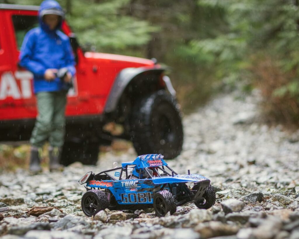 boy in blue rain jacket standing beside red jeep playing with a blue RC car on the rocks